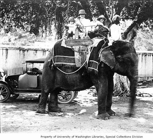 John Dutton Wright, his wife Ysabel Wright, and their children John Jr. and Anna seated on an elephant, with a car in the background, ca. 1921