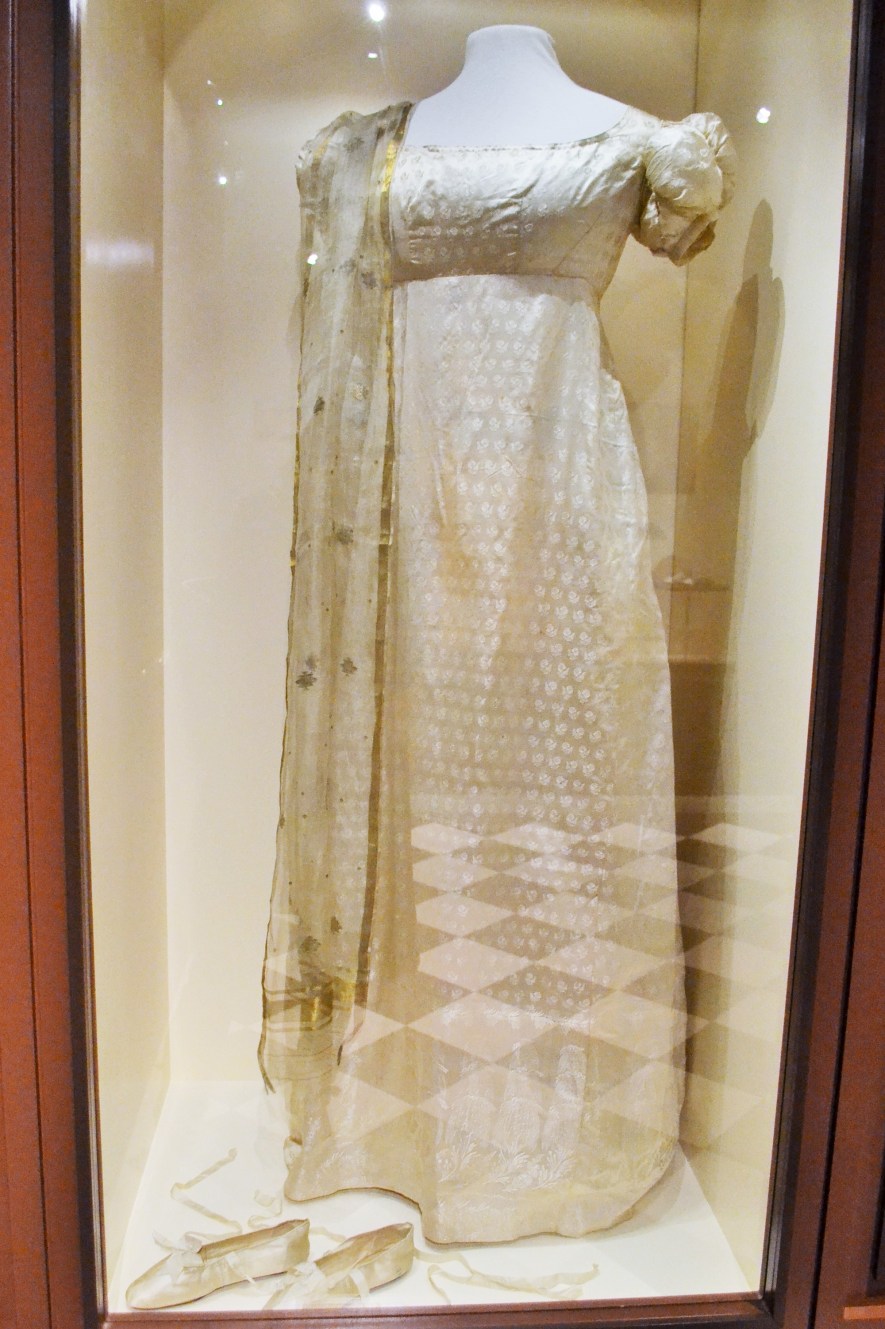 One trend for fashionable women of the 19th century was to dress like Greek goddesses. Their light, gauzy fabrics of cotton muslins, thin luminous silks, or translucent tulle, were considered scandalous by the old guard. Since this trend exposed bosoms and arms, young women supposedly succumbed to “muslin fever” from being underdressed for cold weather!  Dress: American cream silk, c.1814 (Collection of Bata Shoe Museum); worn with shawl of Benares silk from India, early 1800s (Private Collection).  Shoes: French, c.1815-1820 (Collection of Bata Shoe Museum) 